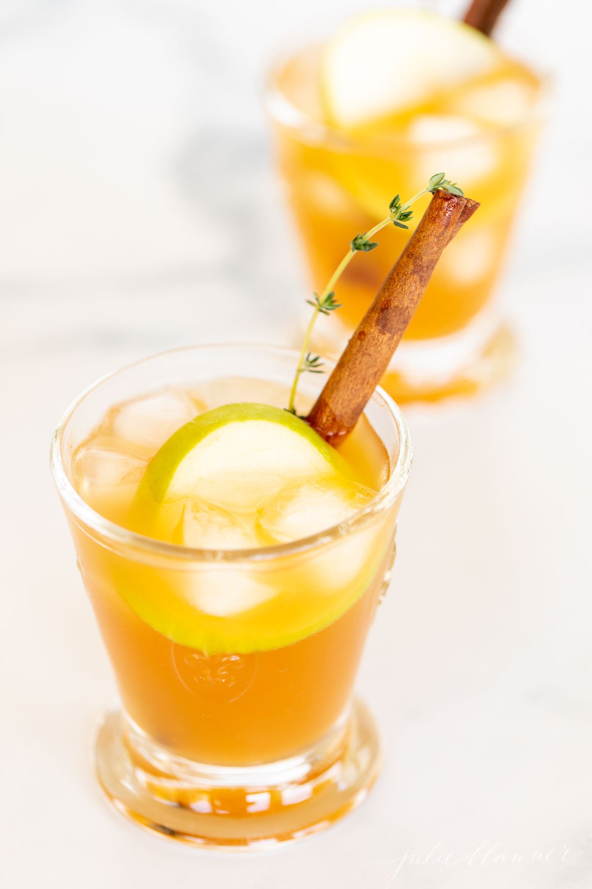 An apple cider cocktail on a white marble countertop, garnished with an apple slice and cinnamon stick.