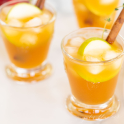Three glasses filled with apple cider cocktails, garnished with apple slices and a cinnamon stick.