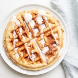 A Belgian waffle topped with creme fraiche drizzle, on a white plate with a linen napkin to the side.