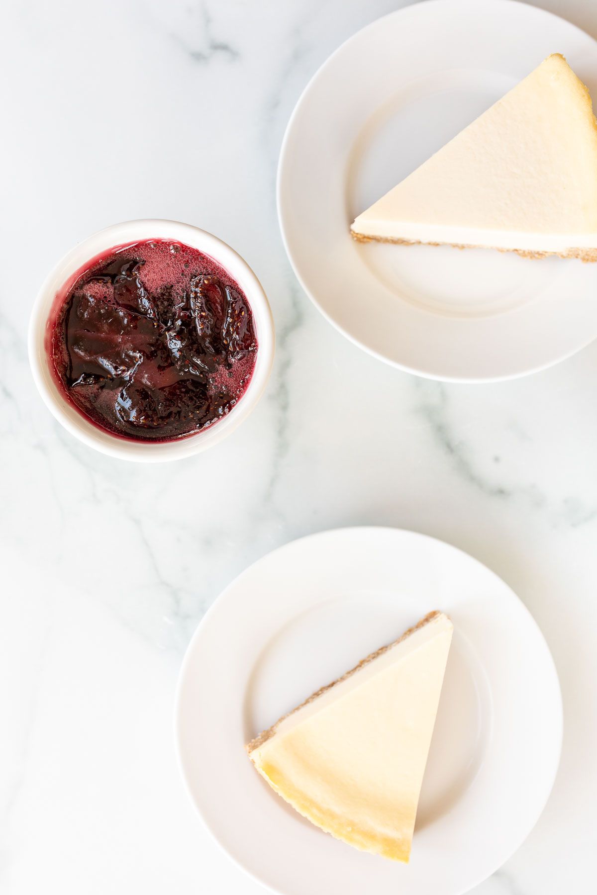 Two plates with a slice of cheesecake, a bowl of strawberry topping in between