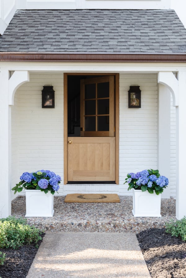 A white brick house with a wooden Dutch door and white planters on the spring porch, filled with blue hydrangeas.