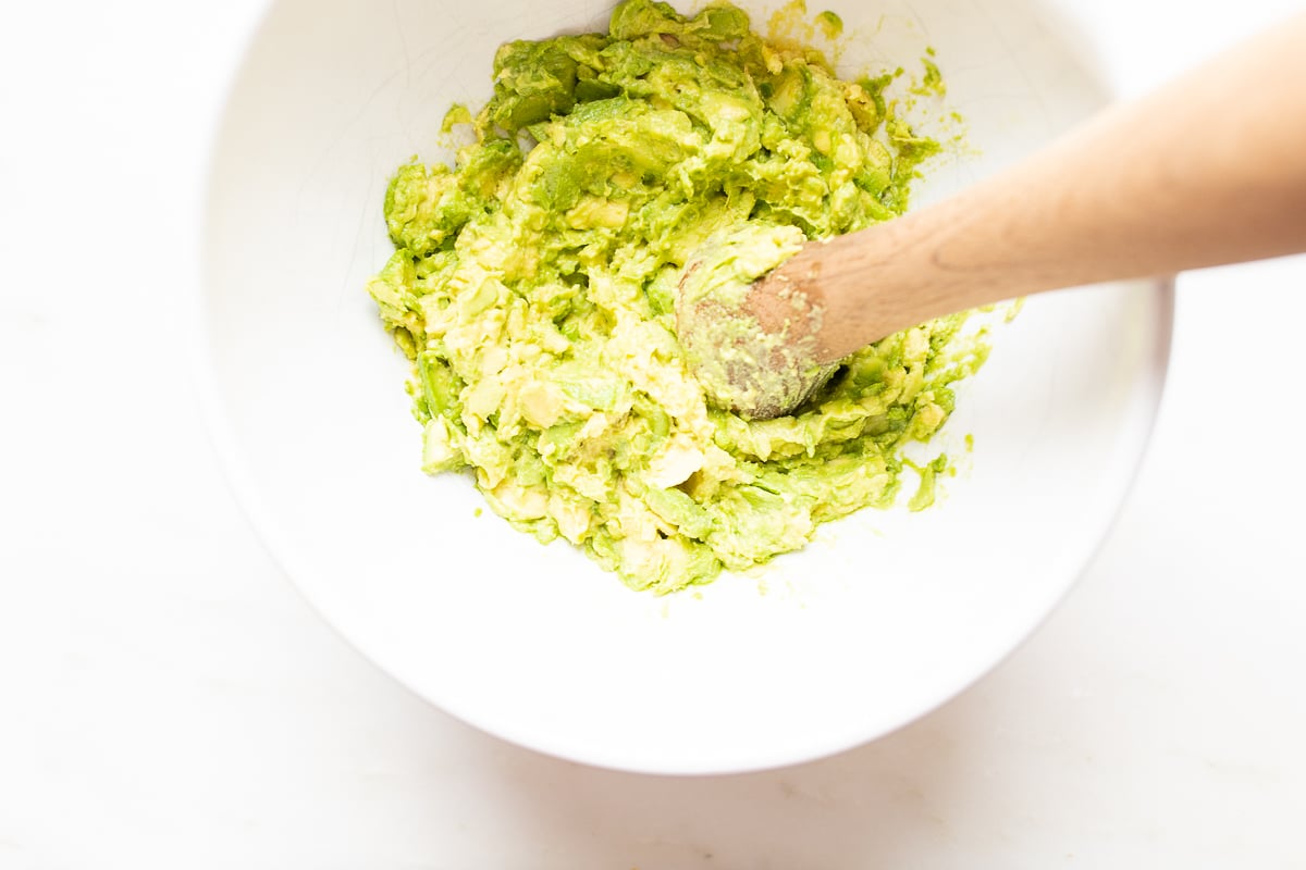 A wooden pestle mashing avocados in a white bowl.