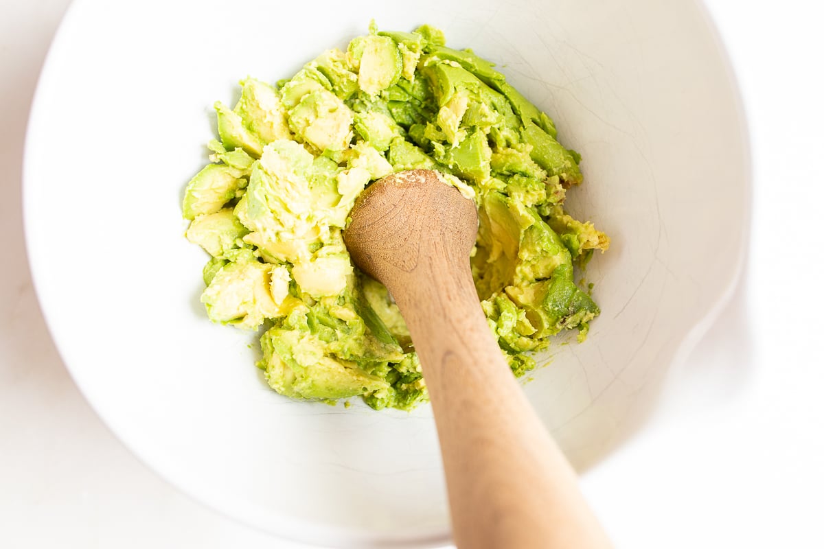 A wooden pestle mashing avocados in a white bowl.