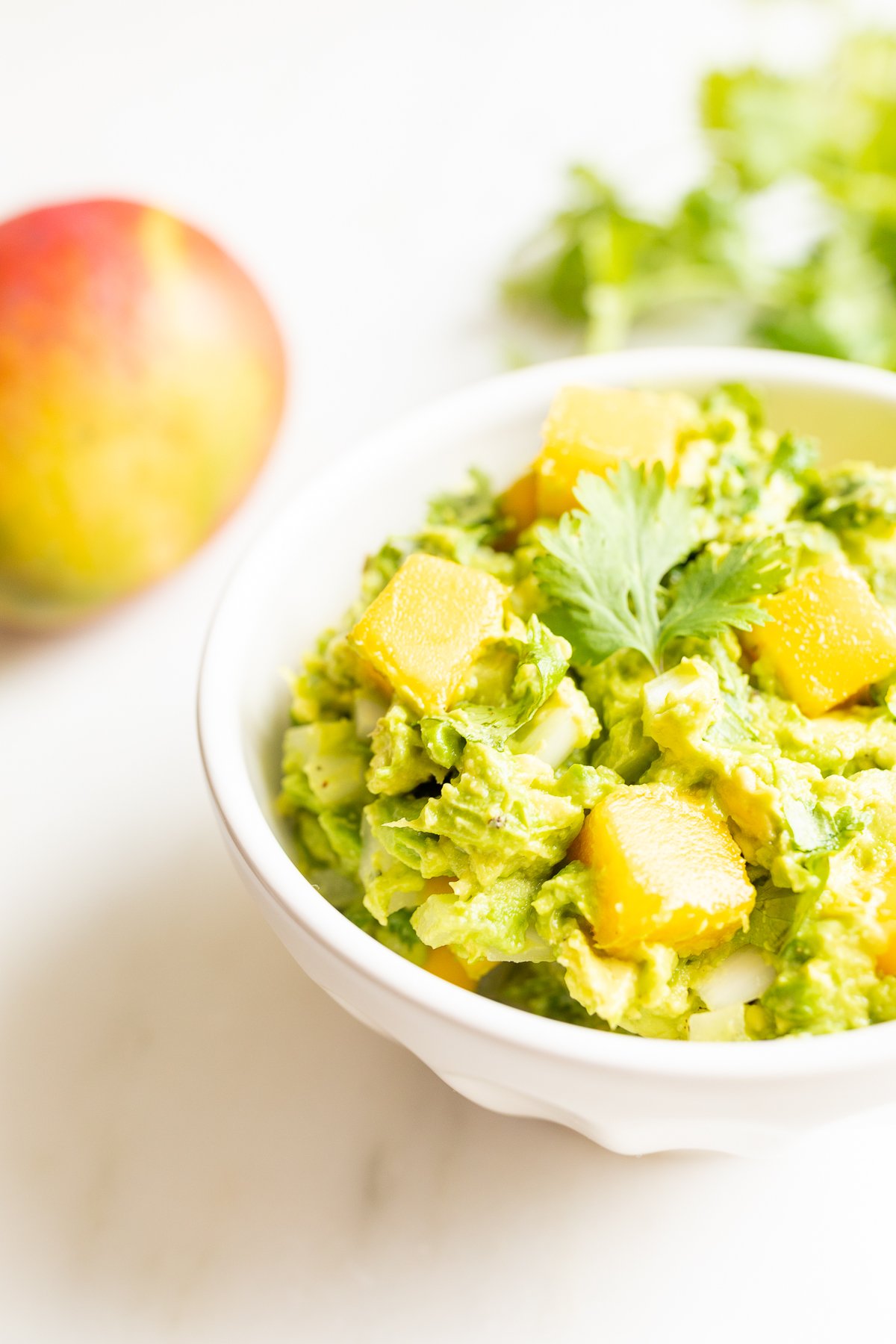 A bowl of mango guacamole surrounded by a mango and fresh cilantro to the side. 