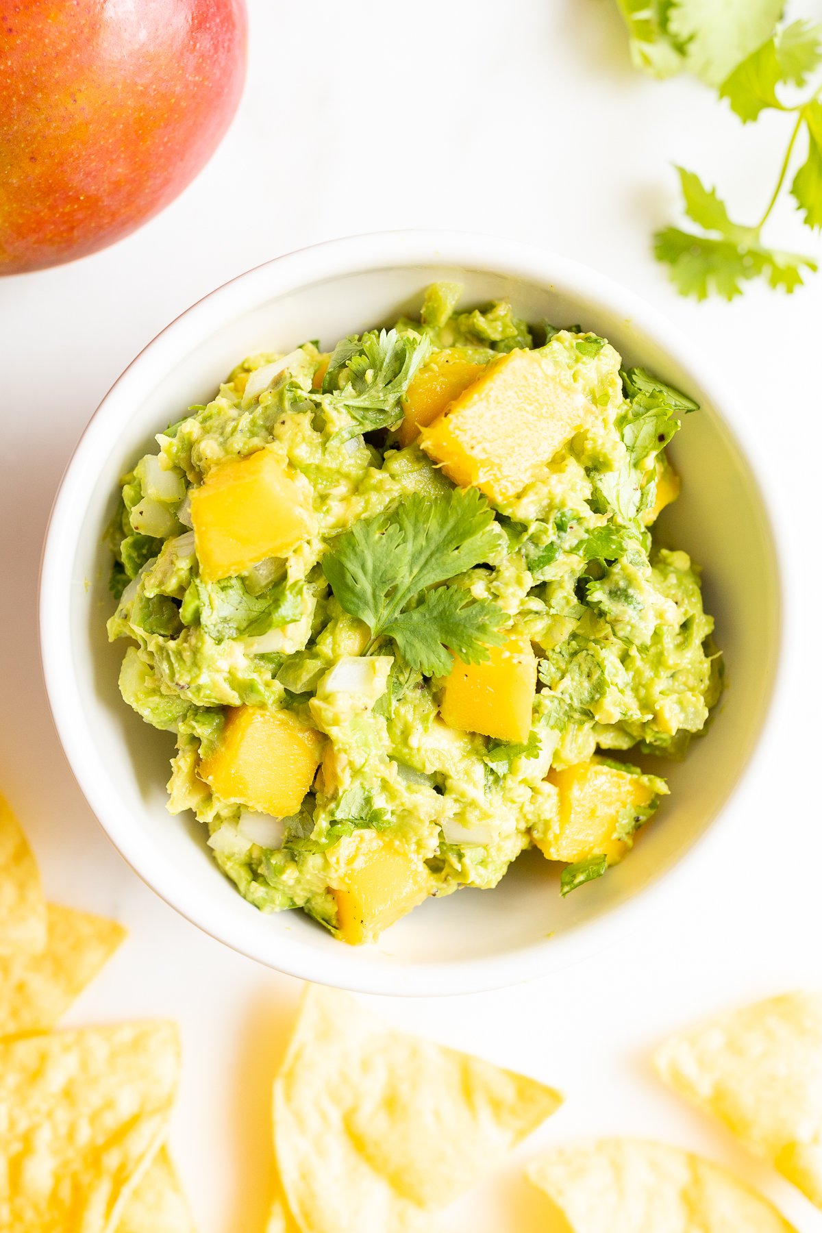 A bowl of mango guacamole surrounded by tortilla chips, along with a mango and fresh cilantro to the side.