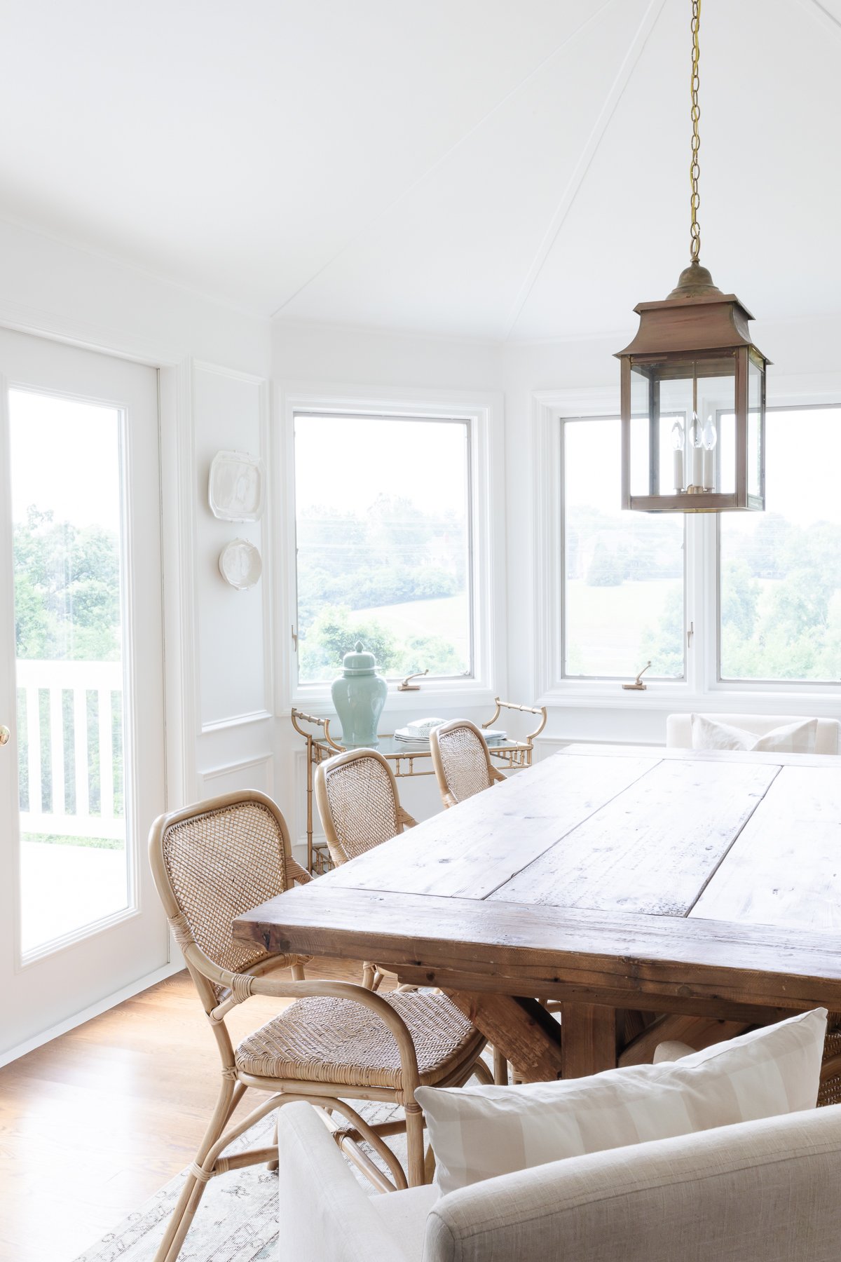 A white dining room with a wood farm table, rattan chairs, brass lantern and plates on the wall.