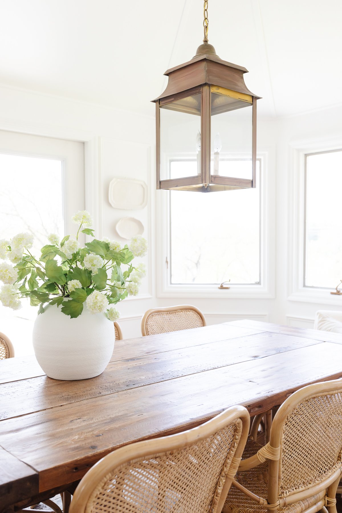 A white dining room with a wood farm table, rattan chairs, brass lantern and plates on the wall.