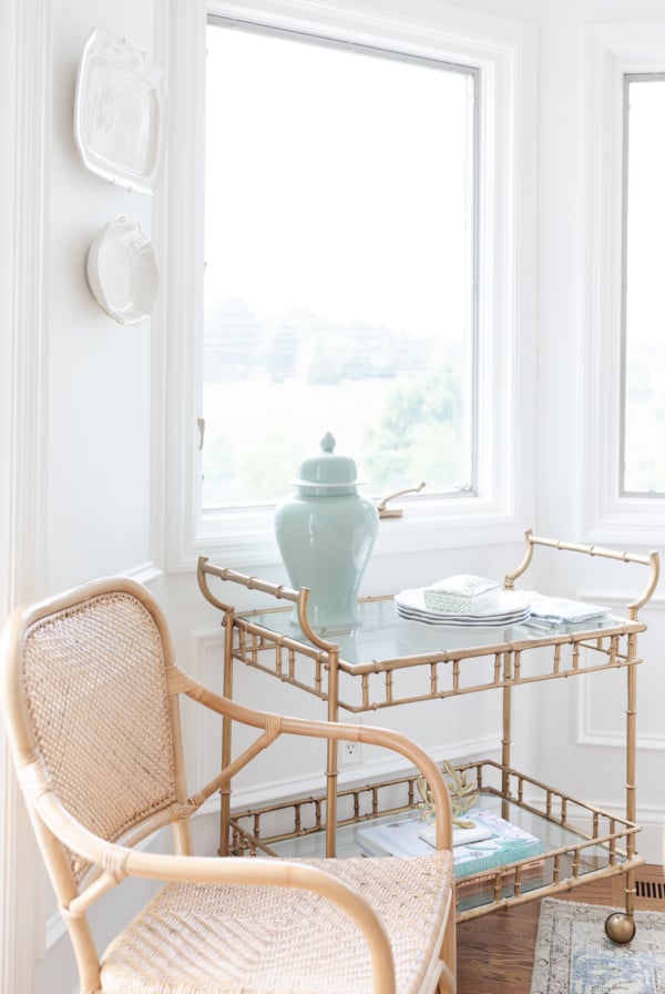 A gold bar cart styled with blue and white accessories in a white dining room. Rattan chair nearby.