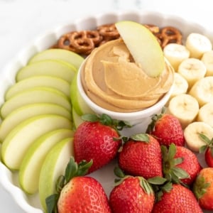 A white platter, with peanut butter dip in the center, surrounded by fruit and pretzels.