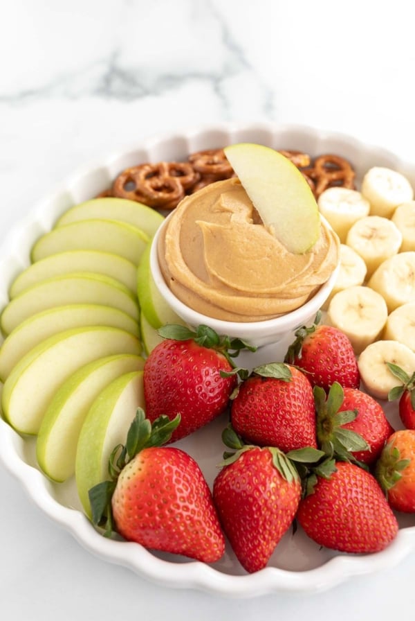 A white platter, with peanut butter dip in the center, surrounded by fruit and pretzels.