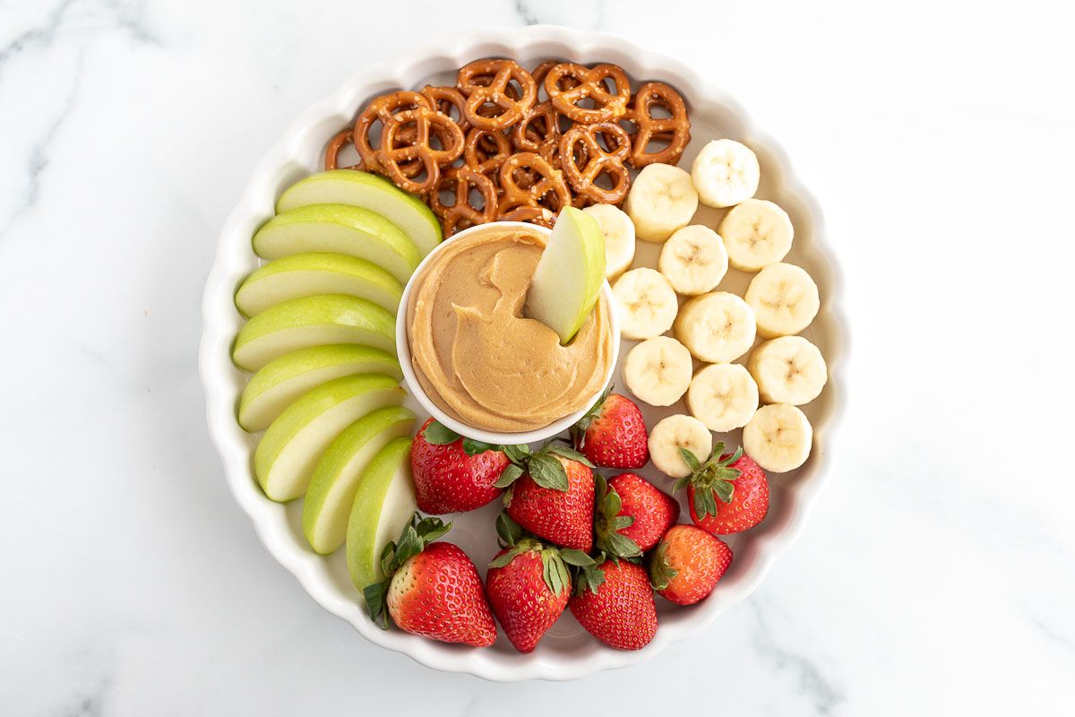 A white platter, with peanut butter dip in the center, surrounded by fruit and pretzels.