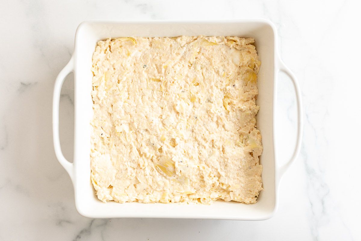 A square white baking dish filled with an artichoke dip recipe prior to baking