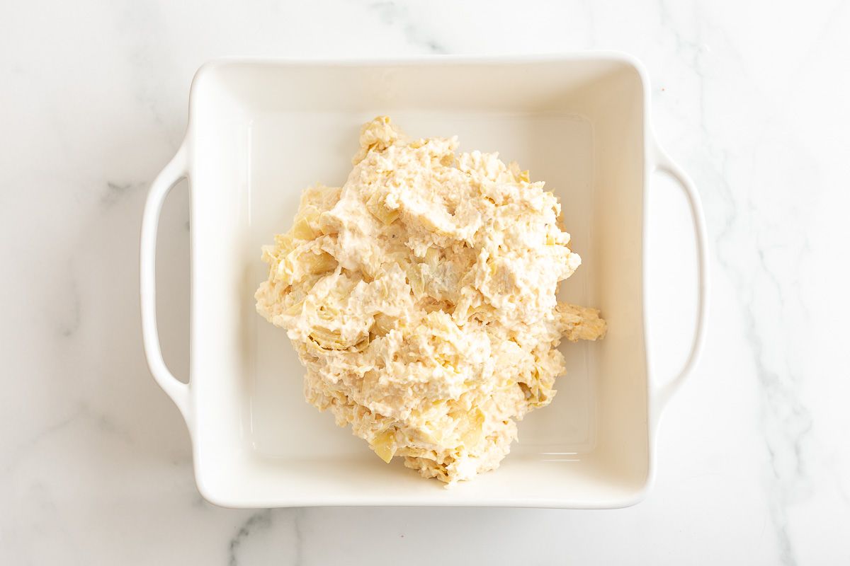 A square white baking dish filled with an artichoke dip recipe prior to baking