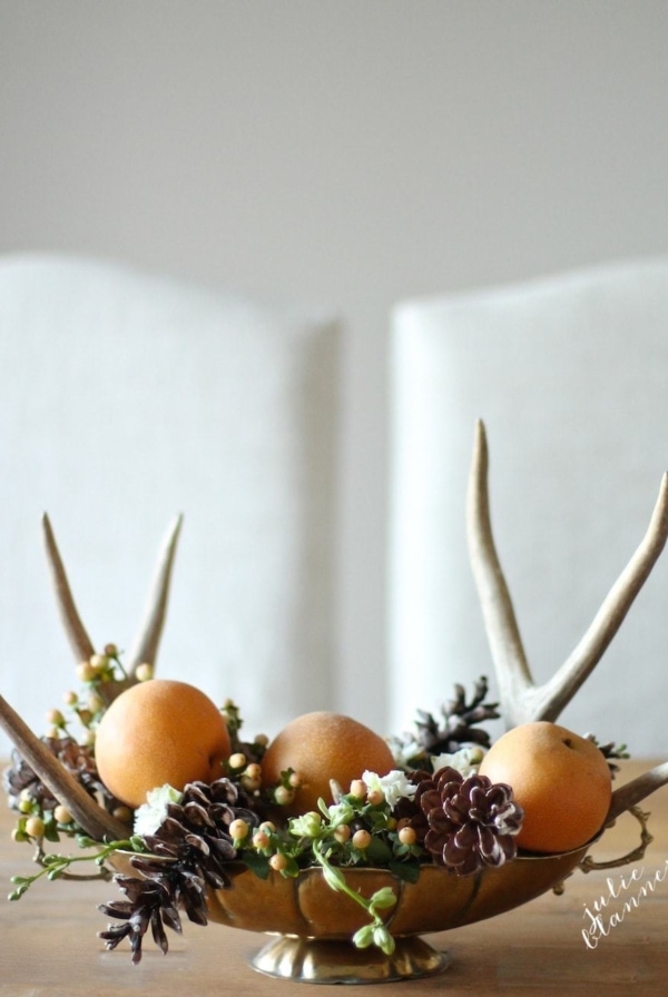 A DIY Thanksgiving centerpiece of a brass bowl full of fruit, pine cones and antlers