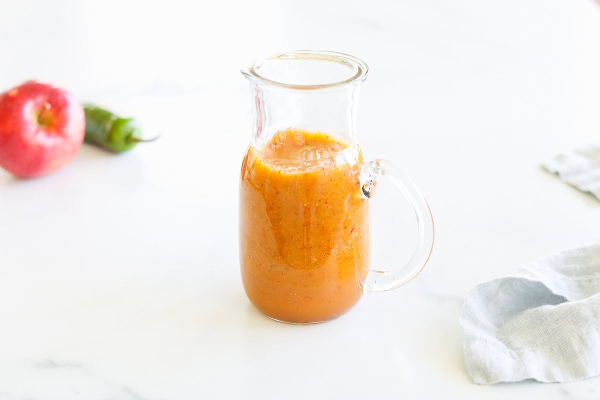 A clear glass pitcher filled with homemade apple smoothie on a white marble countertop, accompanied by a whole apple and a bottle of chipotle salad dressing.