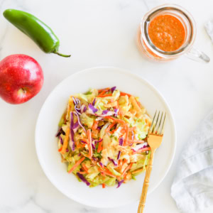 A colorful dish of mixed salad next to an apple, a green chili pepper, and a jar of chipotle salad dressing, all presented on a marble surface.