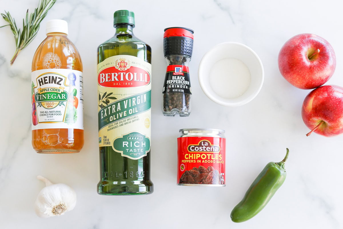 Various ingredients on a marble countertop, including olive oil, vinegar, black pepper, chipotle vinaigrette, a jalapeño, garlic, apples, and rosemary.