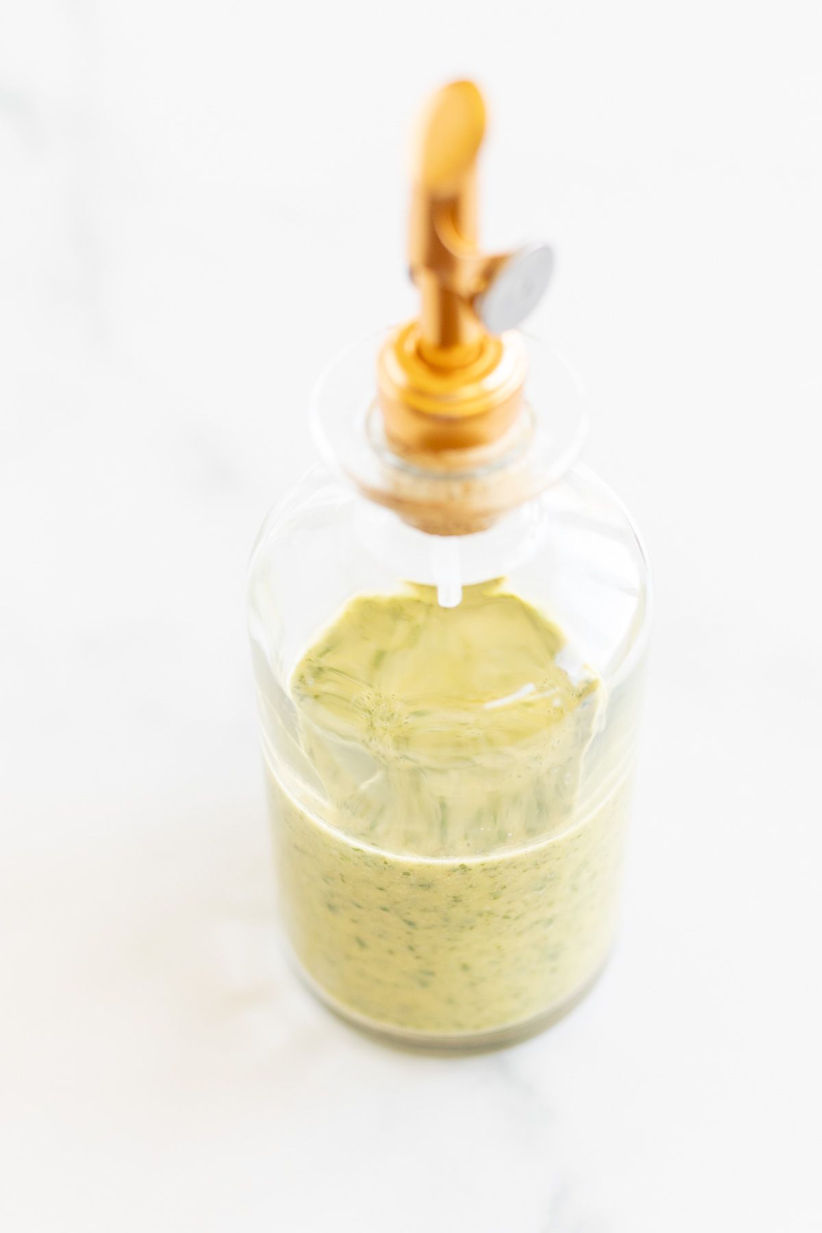 A glass bottle with a gold spout, full of cilantro lime dressing. Placed on a white marble countertop.