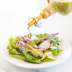 A glass bottle of cilantro lime dressing being poured over a green salad on a white plate.