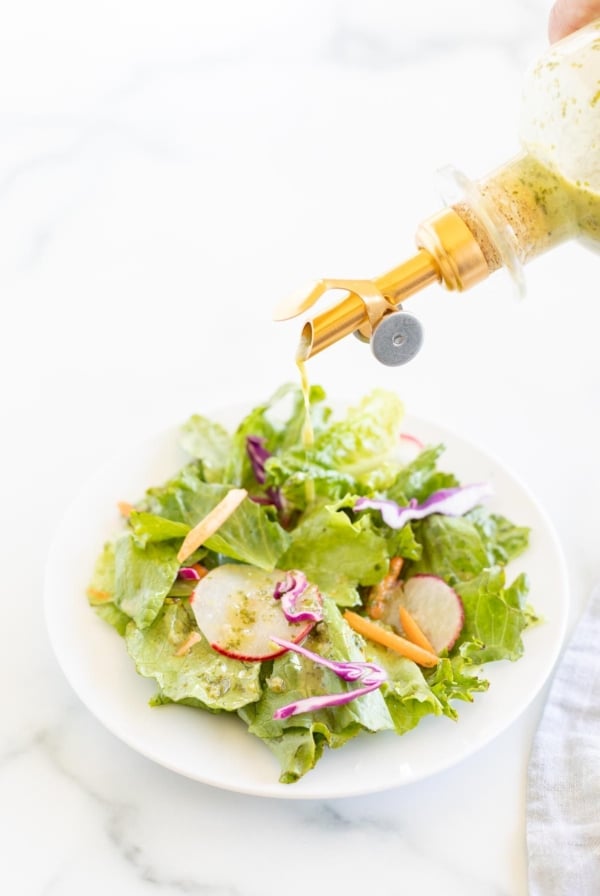 A glass bottle of cilantro lime dressing being poured over a green salad on a white plate.