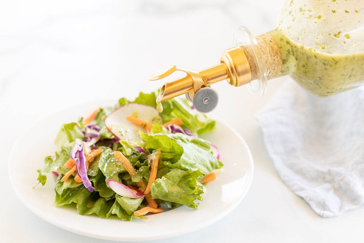 A glass bottle of cilantro lime dressing being poured over a green salad on a white plate.