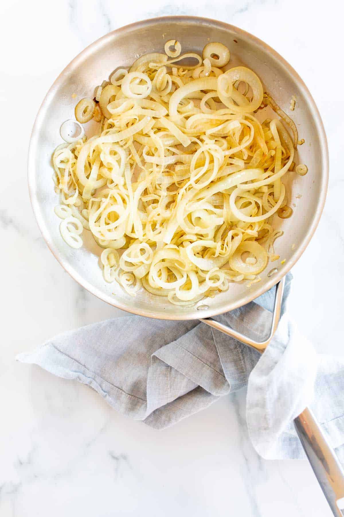 A silver saute pan on a marble surface, filled with caramelized onions