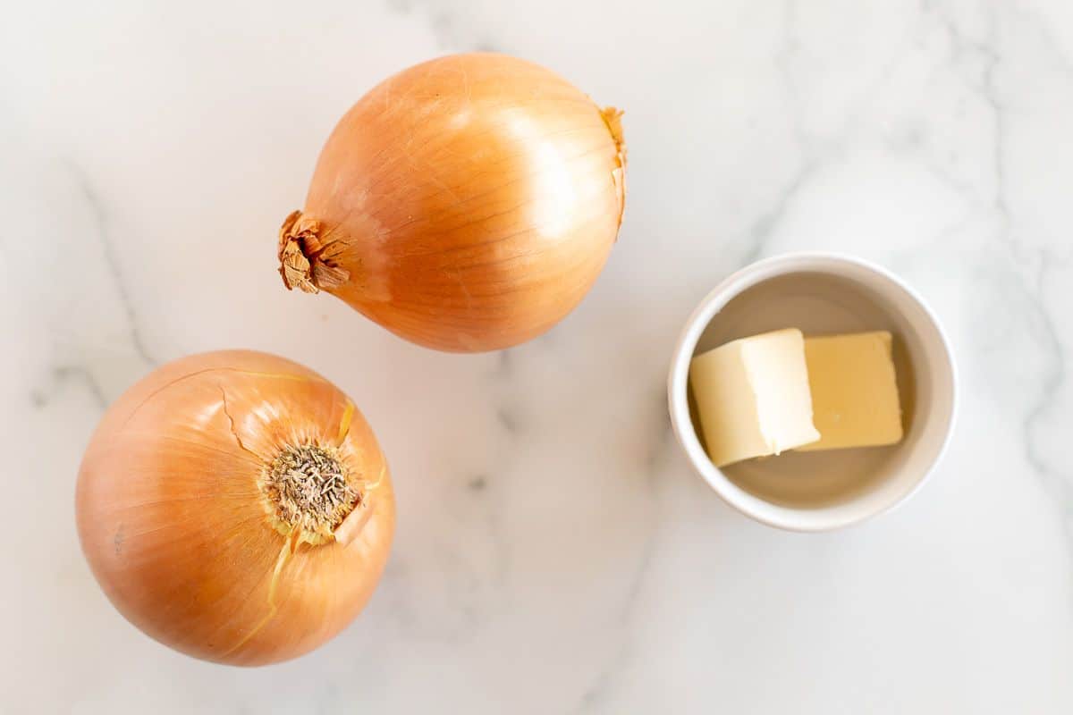 A marble surface topped with two whole yellow onions and two tabs of butter.