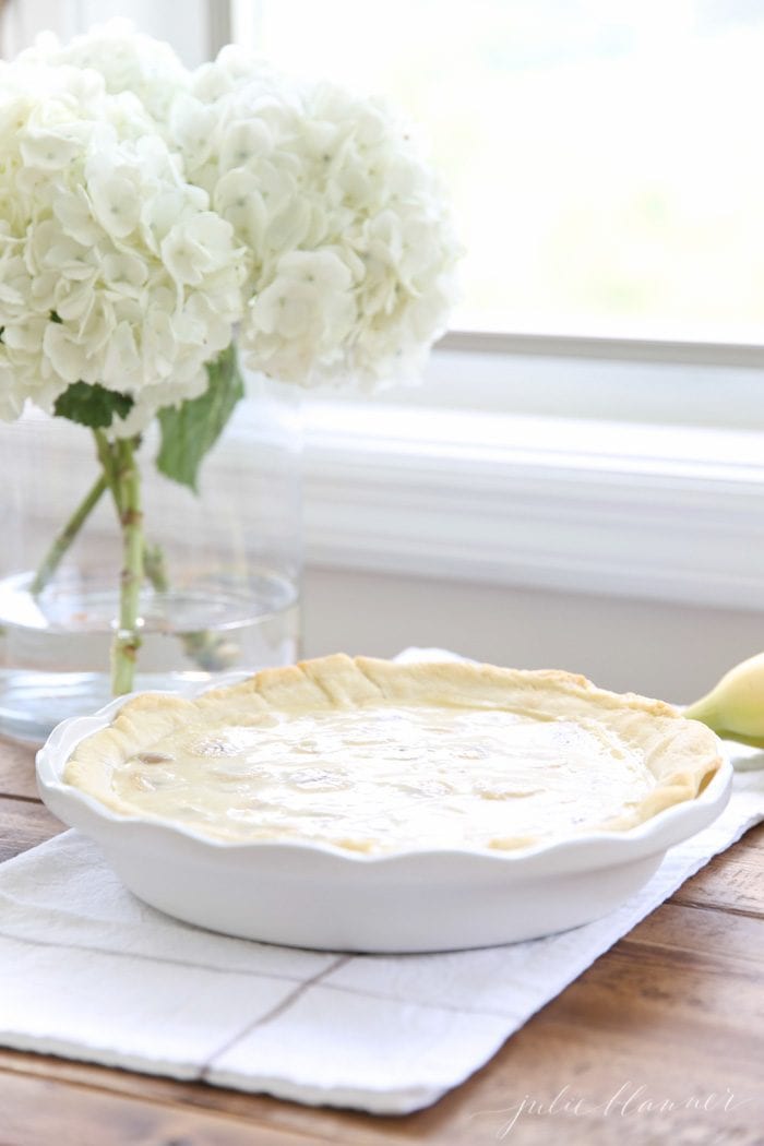 easy banana cream pie recipe in white pie dish with white flowers in vase in background