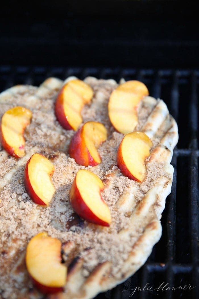 Pizza dough spread out on an outdoor grill, topped with sliced peaches, cinnamon and brown sugar