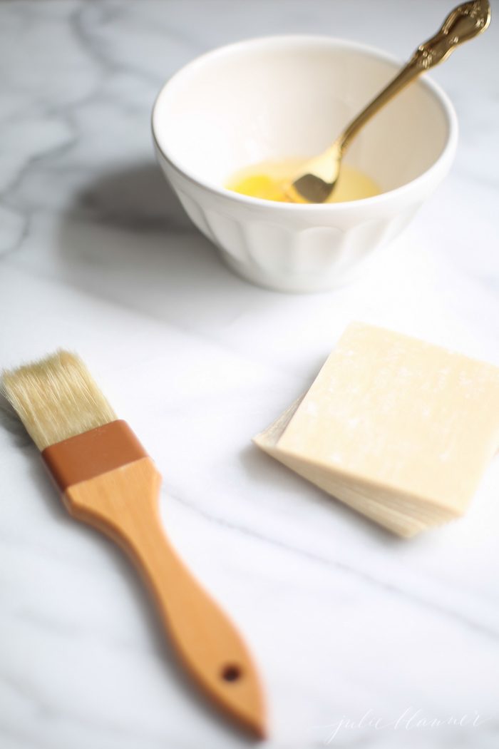 pastry brush, stack of wonton wrappers, and beaten egg with fork in a white bowl