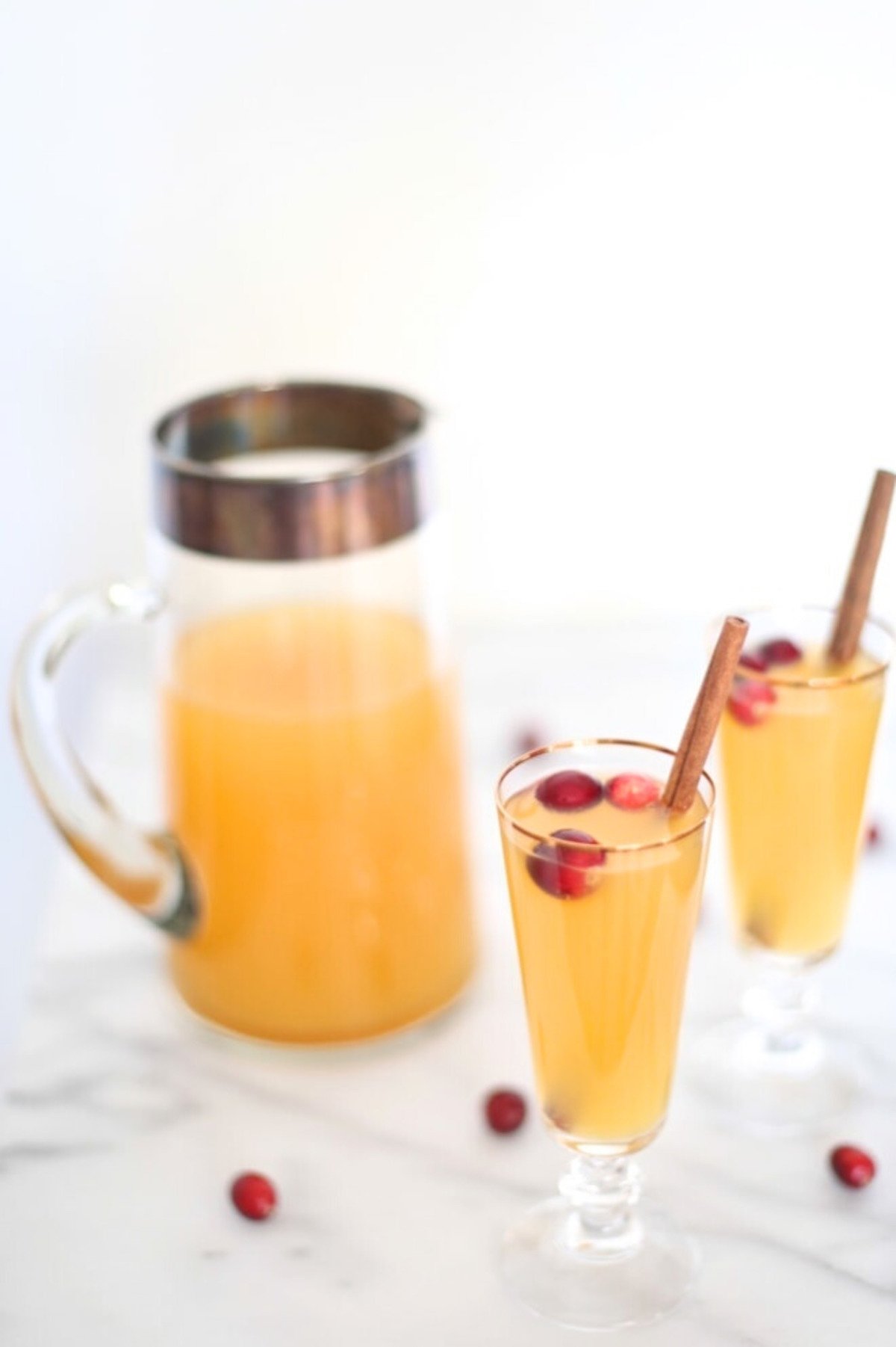 Two glasses of light orange pear prosecco cocktail garnished with cranberries and cinnamon sticks, alongside a glass pitcher containing more of the same drink, set on a white marble surface.