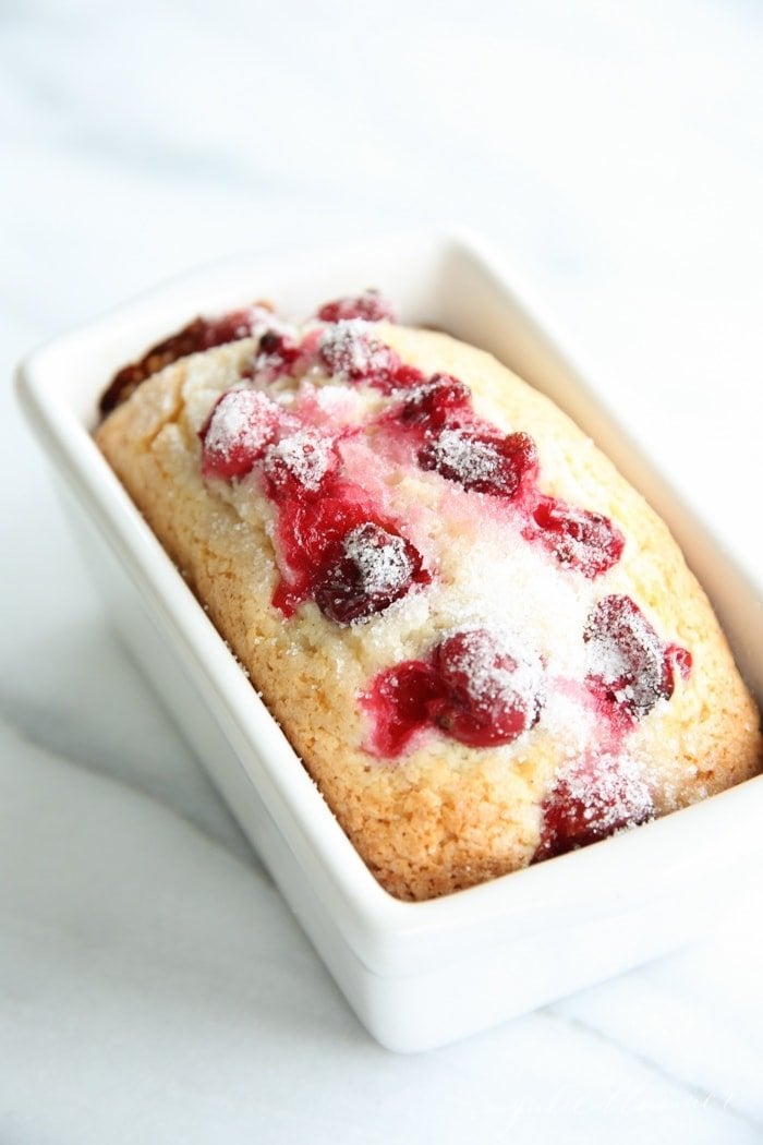 A small loaf of bread with cranberries on top, in a white ceramic loaf pan, white background.