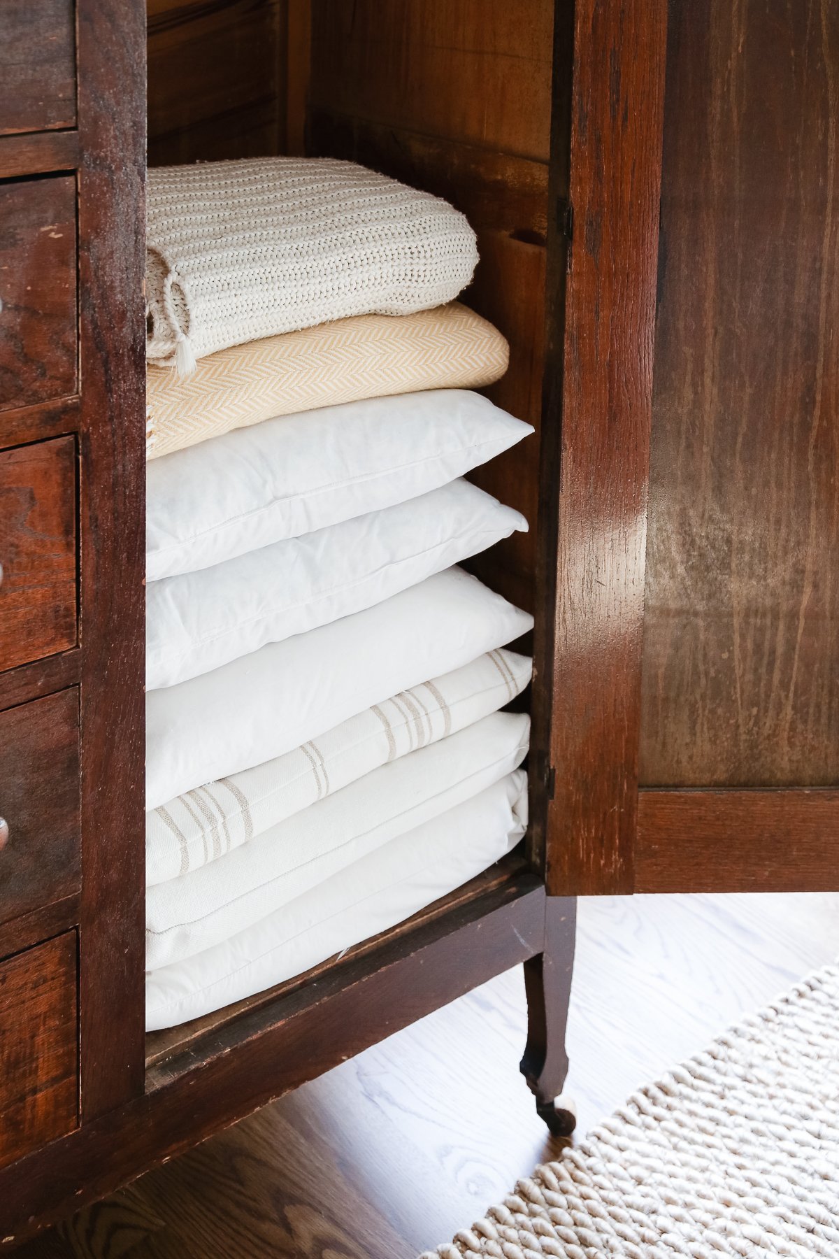 A dark wood linen cabinet in a family room space, with the door opened to reveal the interior storage.