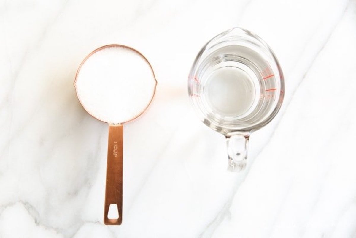 A measuring spoon with white powder next to a glass measuring cup of simple syrup on a marble surface.