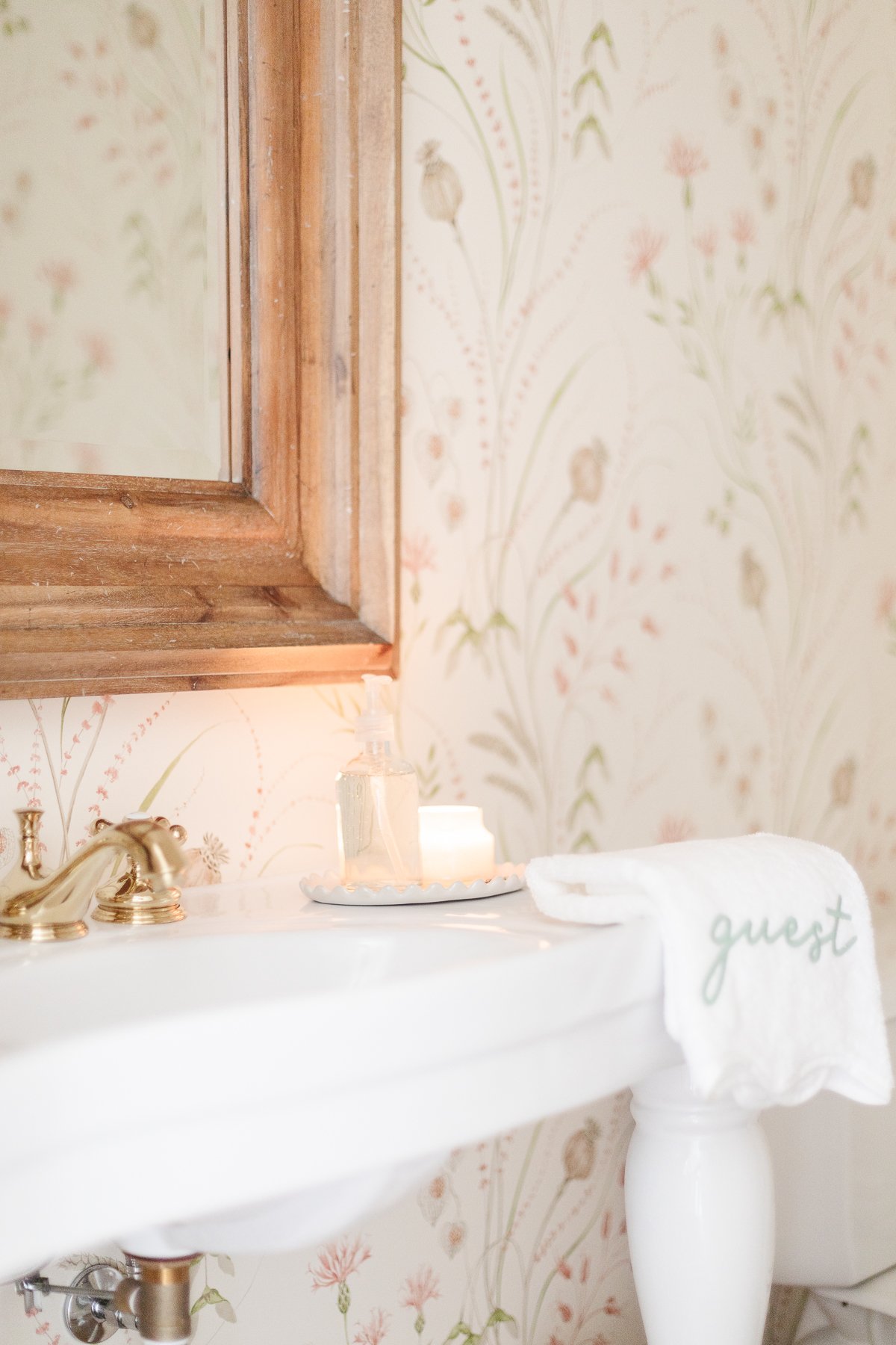 A wallpapered powder room with a wood mirror and white pedestal sink.