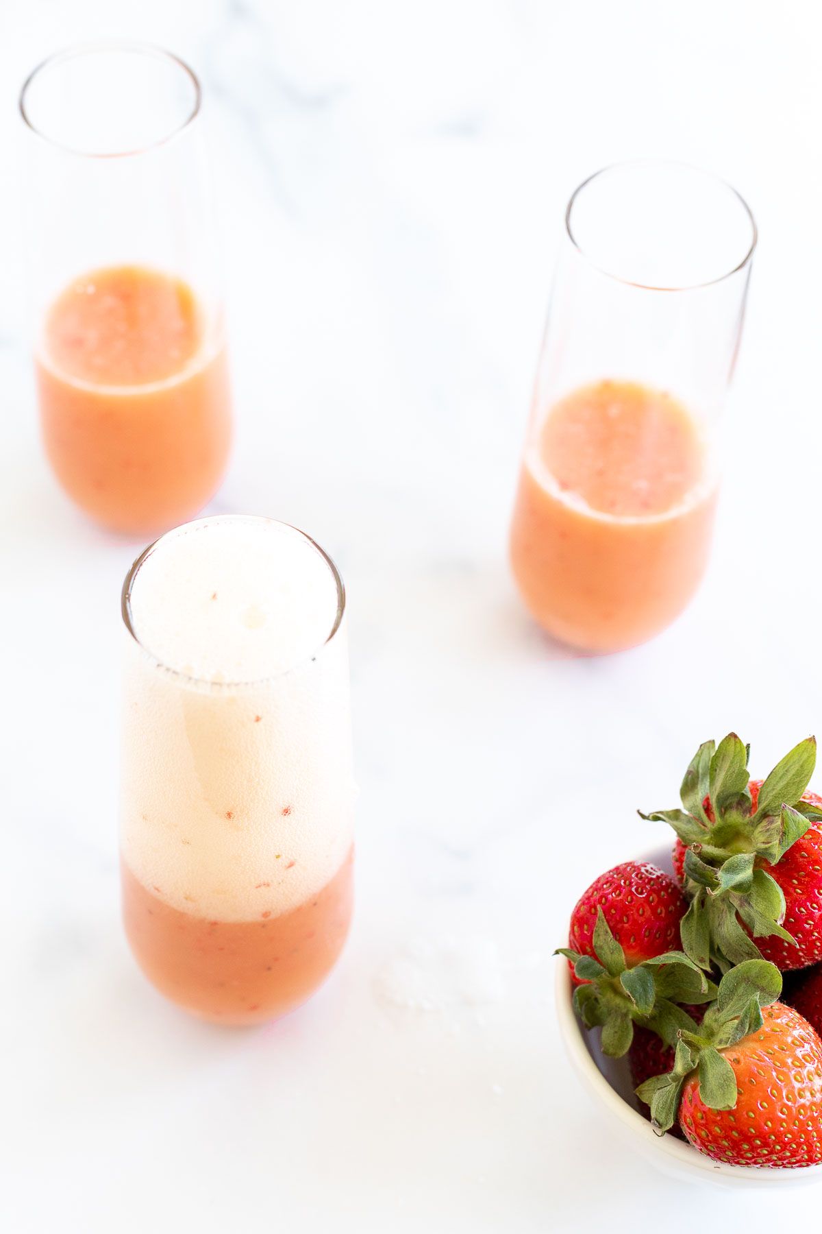 A bottle of Champagne pouring into strawberry mimosas, with a bowl of strawberries on the side