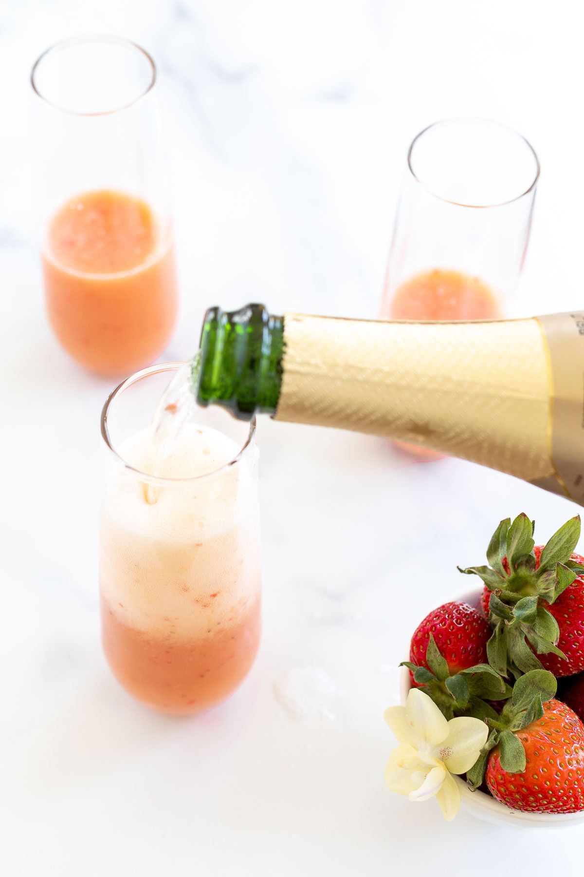 A bottle of Champagne pouring into strawberry mimosas, with a bowl of strawberries on the side