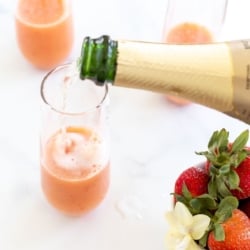 A bottle of Champagne pouring into strawberry mimosas, with a bowl of strawberries on the side