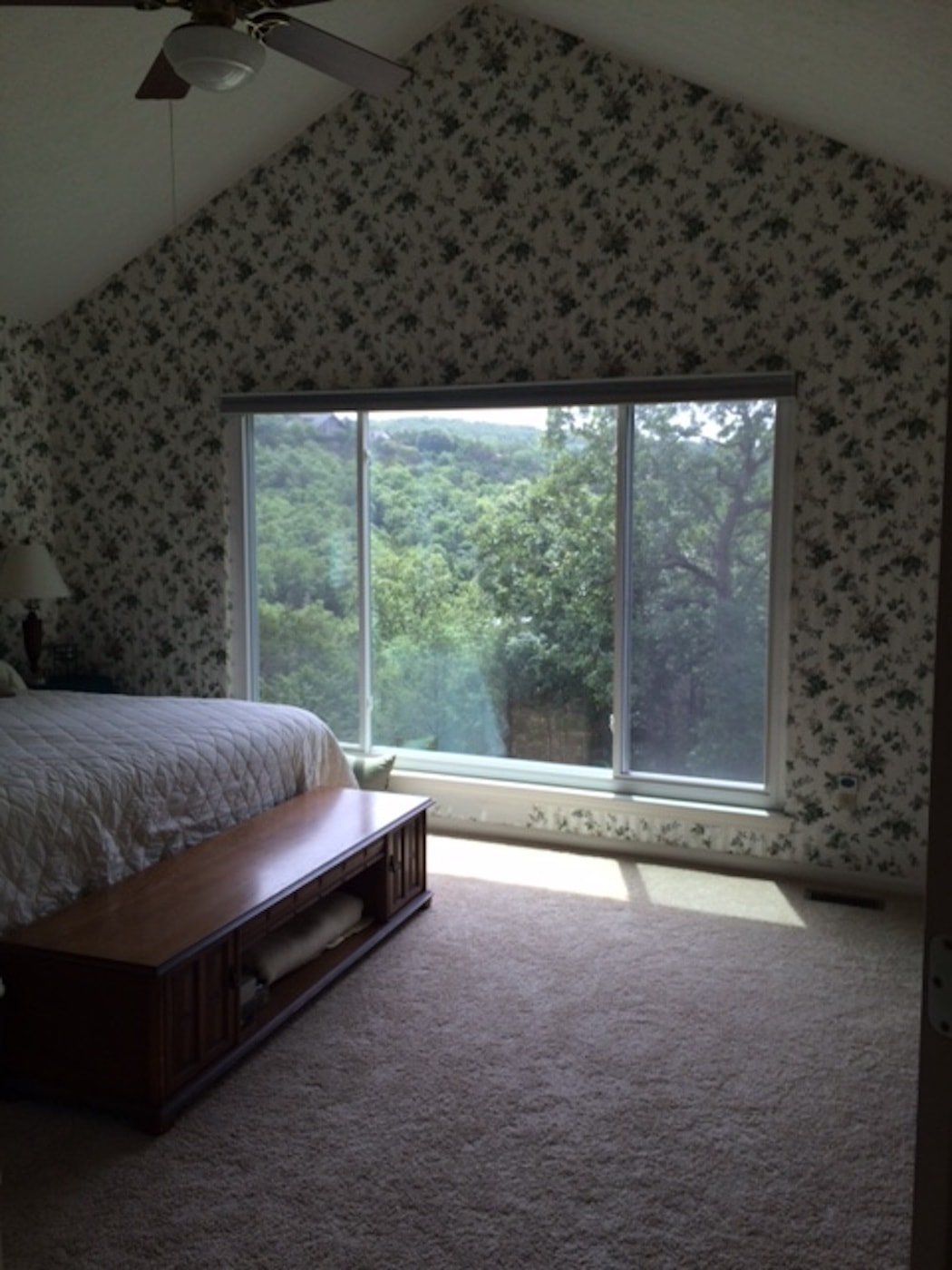 A dark bedroom with dated wallpaper on the walls a large windows.