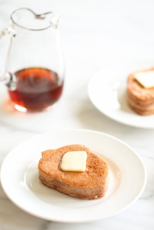 Two white plates each hold a tree-shaped chocolate protein pancake topped with a pat of butter. A glass pitcher of syrup is in the background on a marble surface.