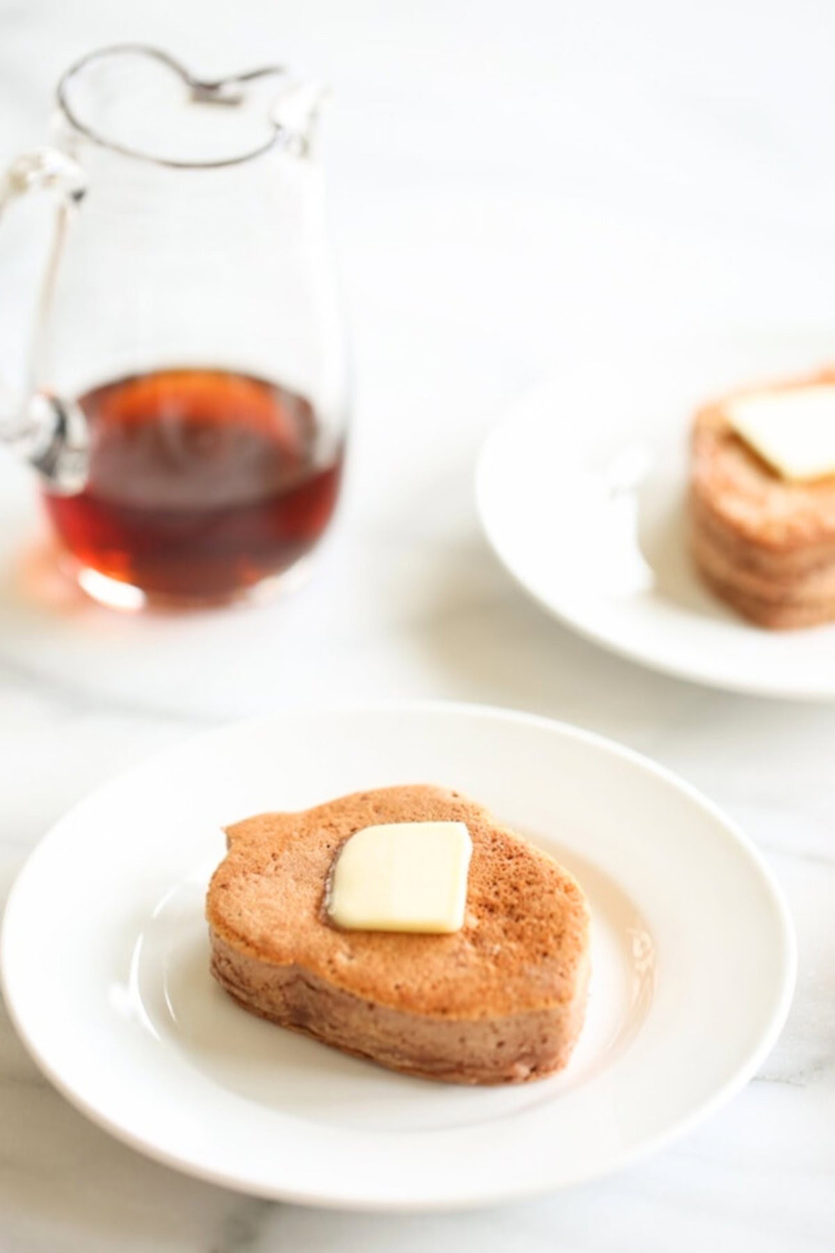 Two white plates each hold a tree-shaped chocolate protein pancake topped with a pat of butter. A glass pitcher of syrup is in the background on a marble surface.