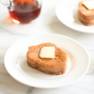 A leaf-shaped chocolate protein pancake topped with a slice of butter is on a white plate. A small jug of syrup is in the background.