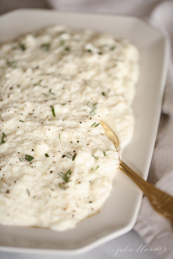 cauliflower mashed potatoes with rosemary in a white platter with gold spoon