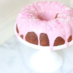 A donut cake on a white pedestal stand, decorated with pink frosting and sprinkles.