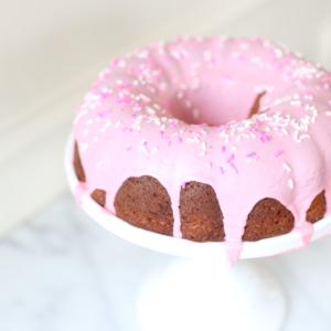 A donut cake on a white pedestal stand, decorated with pink frosting and sprinkles.