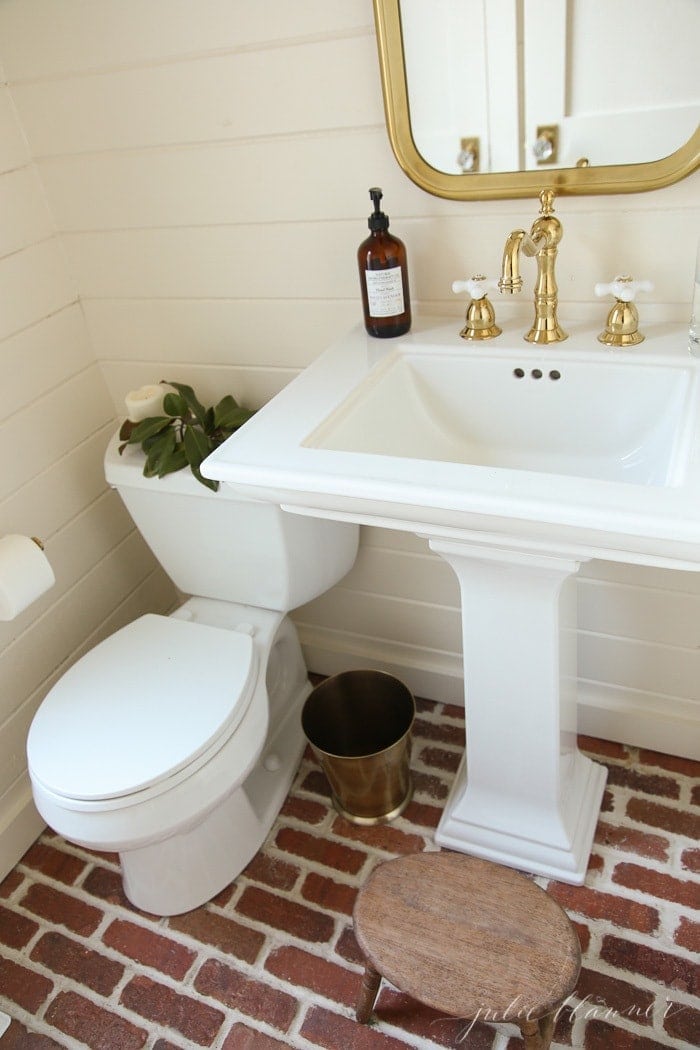 A white bathroom with brick floors, magnolia placed on toilet tank for a little winter decor.