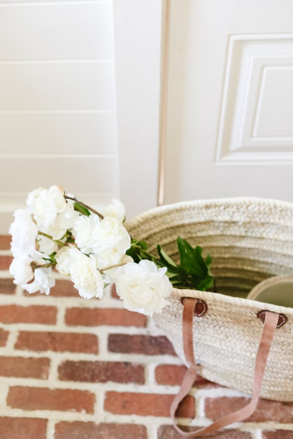 A raffia tote bag full of flowers on red brick flooring.