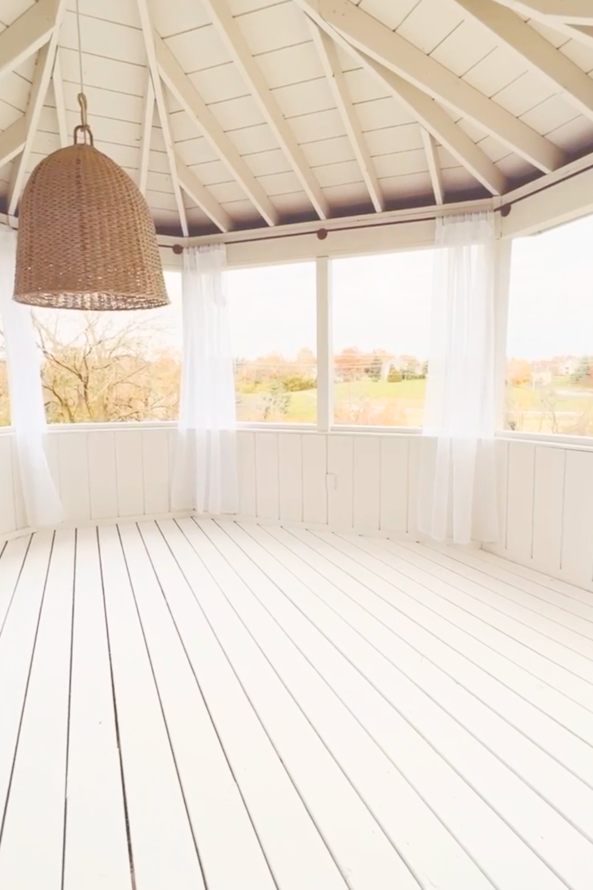 A white, sunlit screened porch with a light wooden floor, sheer white curtains, and a large woven wicker pendant light. Several windows offer a view of a grassy landscape with trees, making it the perfect spot to admire the best deck stain or paint choices for your outdoor space.