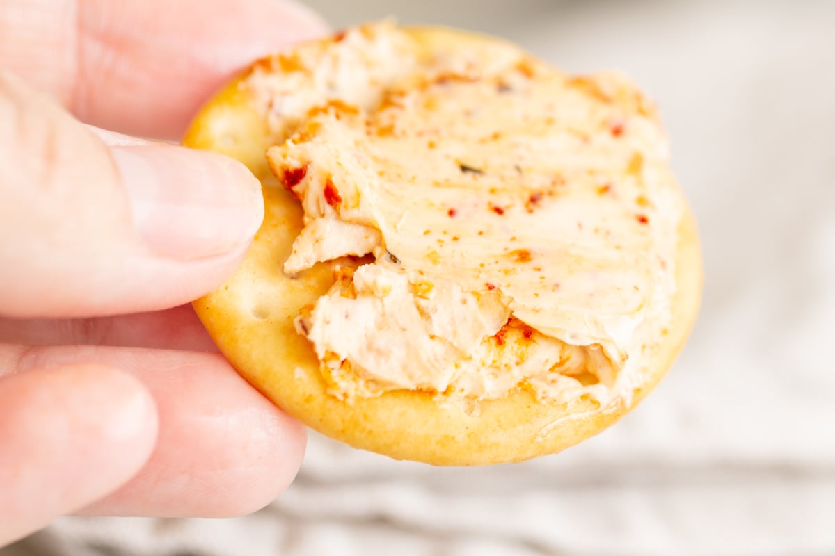 A hand holding a cracker topped with a spread from a spicy cheeseball.