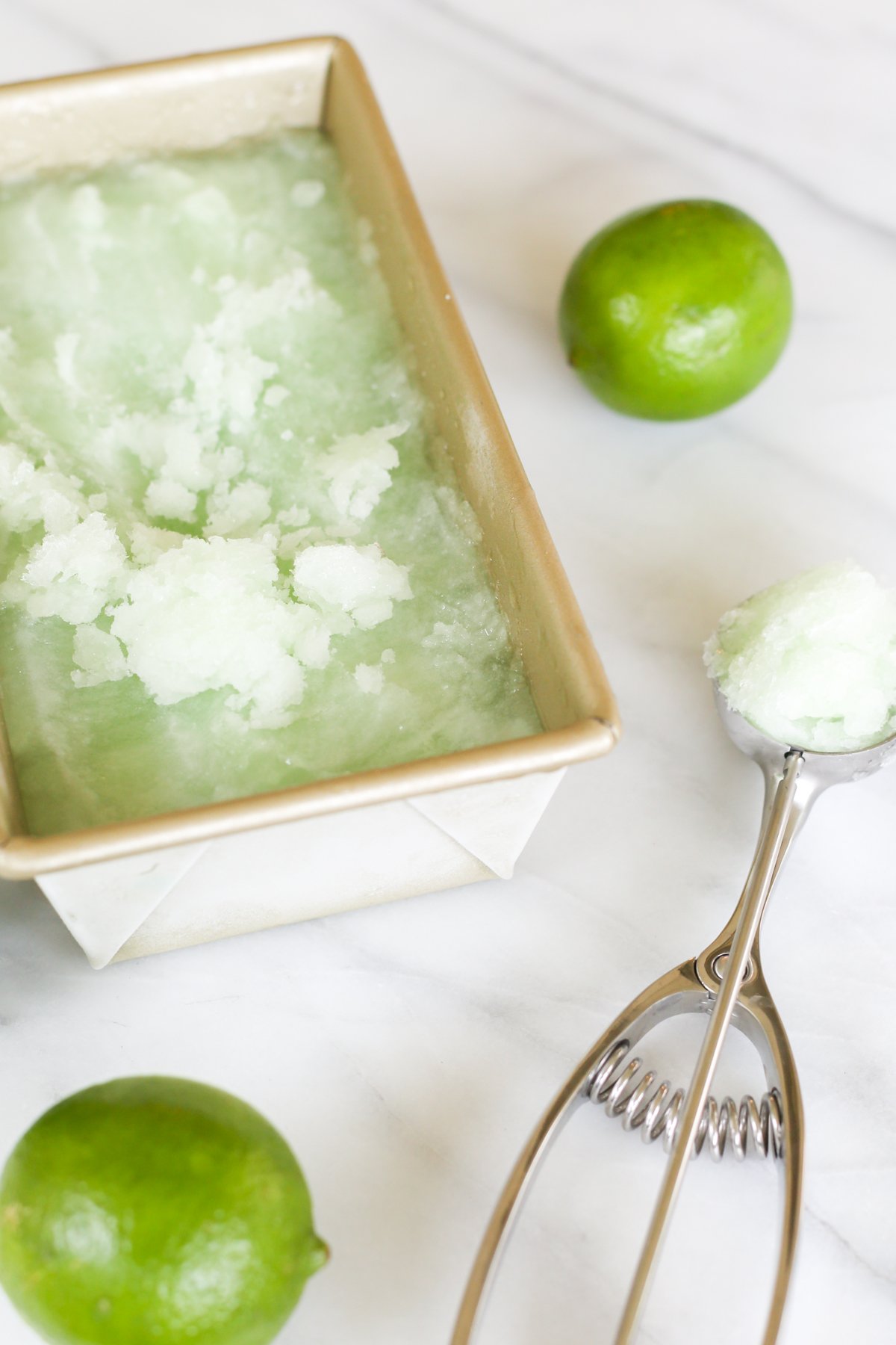 Homemade lime sorbet in a gold loaf pan. A lime and an ice cream scooper are nearby.